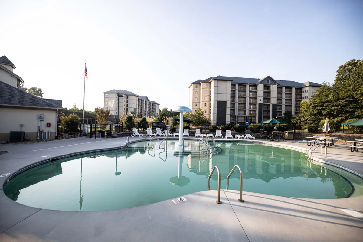 A large swimming pool surrounded by lounge chairs and a playful fountain, with nearby resort buildings in the background. The pool area is lively yet inviting, perfect for families looking to swim, sunbathe, or socialize outdoors.