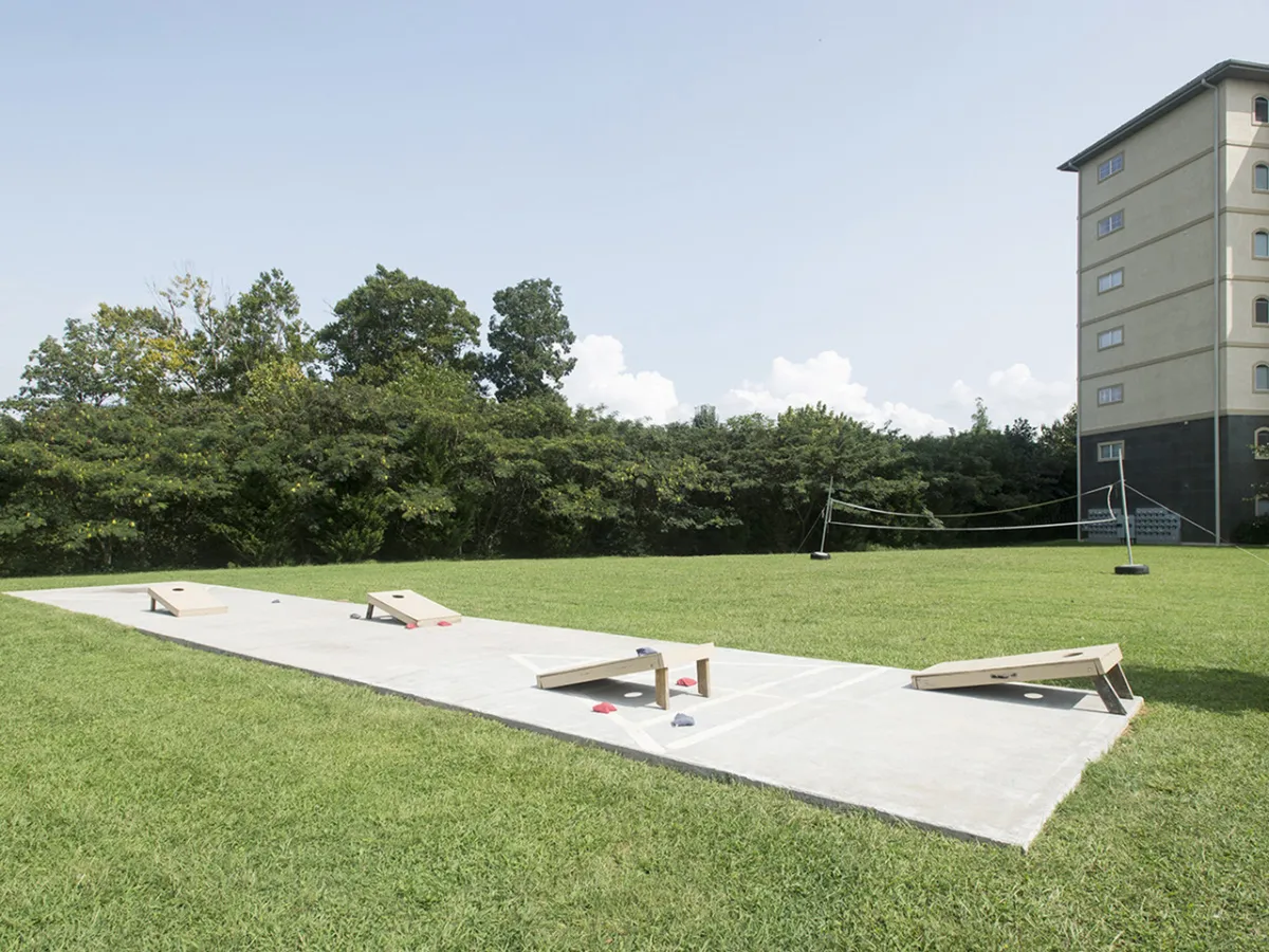 An outdoor scene showing cornhole boards on green grass in front of the condo building, capturing the spirit of social play and group-friendly fun. The background establishes the building's proximity to open communal spaces.