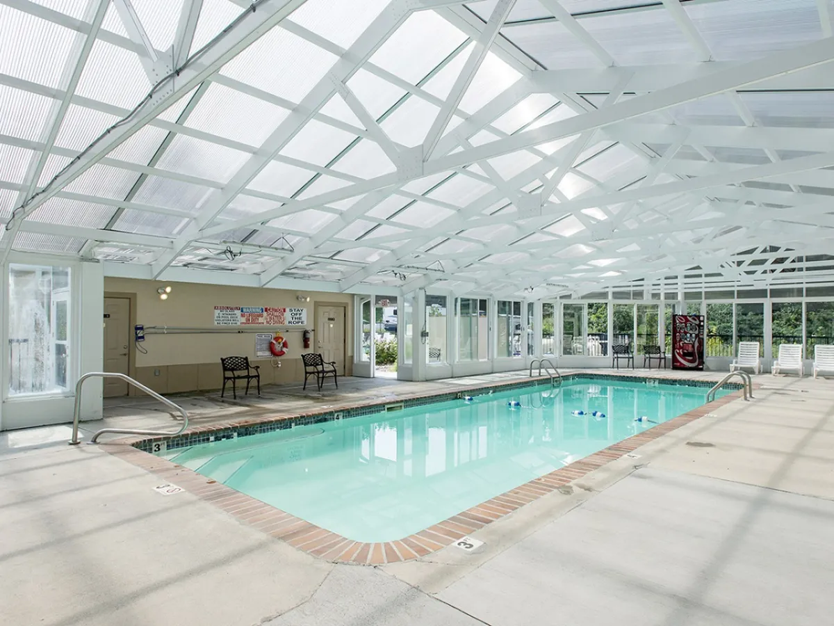 An indoor swimming pool with a transparent roof allowing in natural light. Lounge chairs are arranged along the sides, and safety signs are visible near the water. The mood is cheerful, with ample space for families or groups to relax and swim regardless of season.