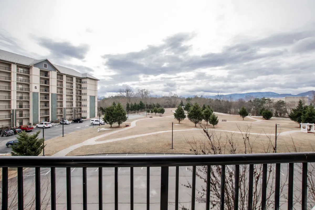 A scenic overlook from a balcony reveals an expansive park area below, framed by trees and backed by distant mountains. The park area invites play, walking, and family gatherings, all set against a beautiful natural backdrop.