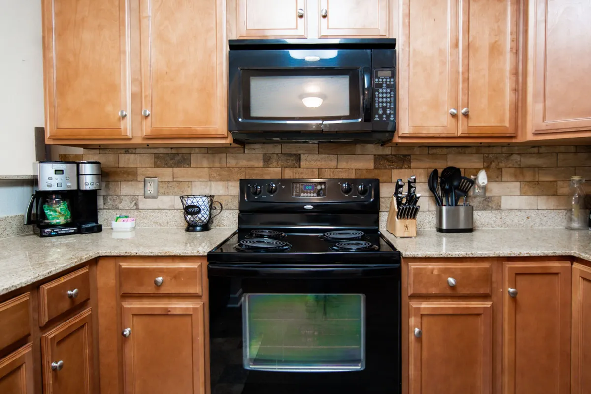 A well-equipped kitchen with a black stove, microwave, granite countertop, and wooden cabinets. The inviting space is ideal for preparing family meals or snacks, underscoring the value and savings of having in-unit amenities.
