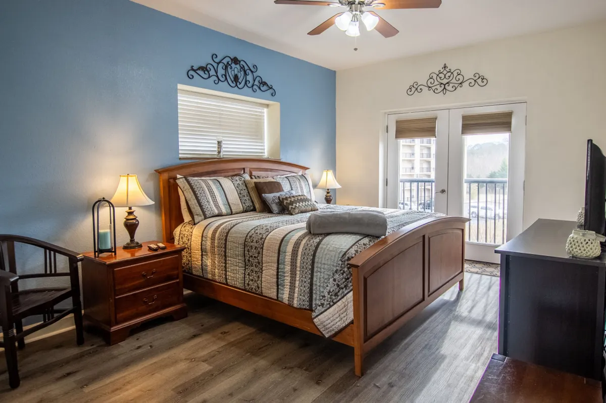 A cozy bedroom with a bed, nightstand, chair, window streaming with natural light, and a TV. The space invites relaxation and quiet moments, catering to various guest needs.