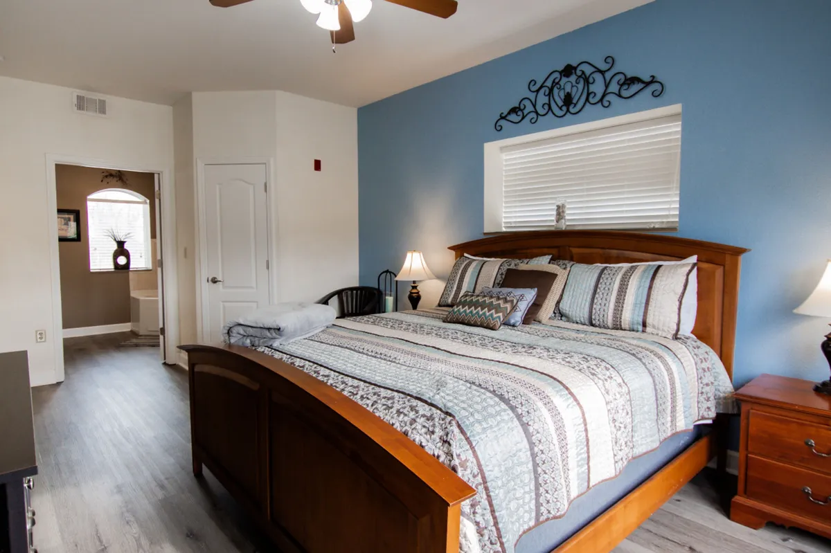 A cozy bedroom with a plush bed, decorative wall art, nightstands with lamps, and warm lighting. The arrangement suggests a restful and inviting space perfect for unwinding after a busy day.