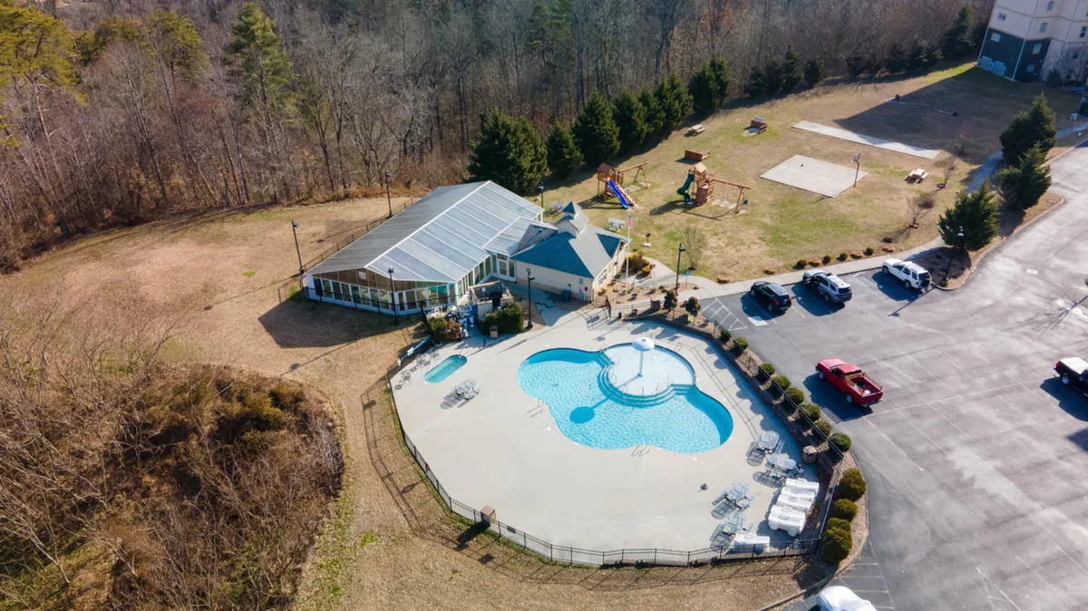 A vibrant outdoor scene featuring a swimming pool, playground equipment, and surrounding trees. The recreation area is adjacent to a parking lot, with children’s play spaces and picnic spots visible, creating a lively, communal environment.