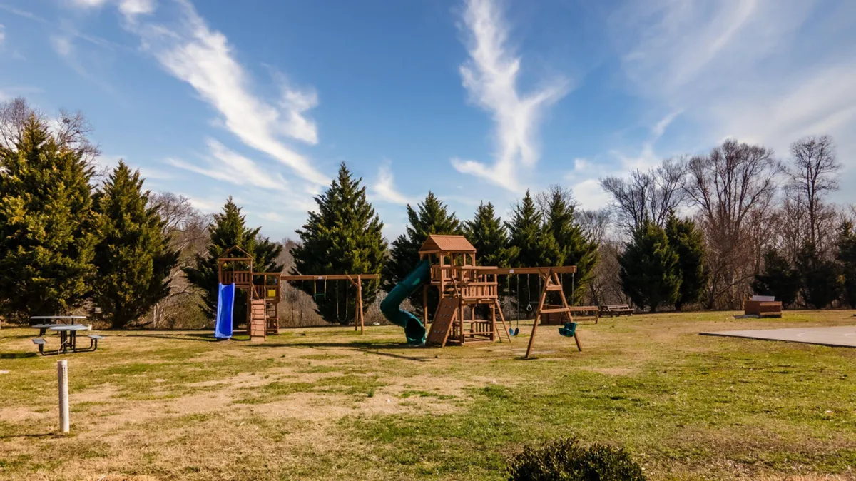 A cheerful playground set with swings, equipment for climbing, and a picnic table, all nestled within green grass and trees. The area exudes joyful energy, perfect for children’s activities and family gatherings outdoors.