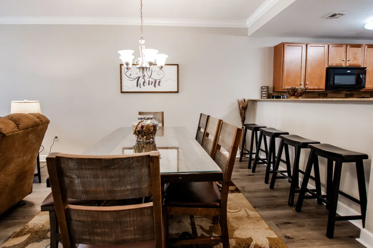 This image captures a spacious open-plan dining and kitchen area. A welcoming dining table sits in the foreground beneath a warm chandelier, with bar stools lining the kitchen's island and wood cabinetry visible in the background. The decor is cozy and homelike, including a framed sign with 'home' over the dining space.